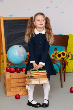 A content girl in a blue school uniform stands and holds a stack of books tied with twine, looking slightly off to the side, in a classroom