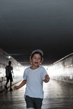 A child in a t-shirt and shorts running down a dark, concrete pedestrian underpass towards the light at the end of the tunnel.