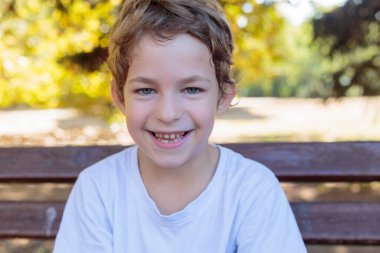 A close-up portrait of a happy, smiling boy with curly hair and sparkling blue eyes, sitting on a bench with a warm, autumn-themed background