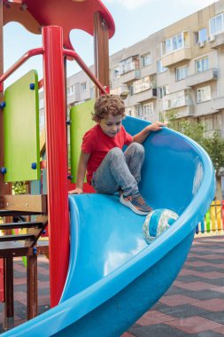 A little boy is sitting on a blue slide at a colorful playground, looking down before sliding