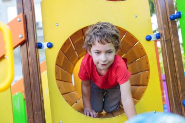 A cute boy is peeking out from a circular window of a playhouse on the playground, smiling and looking at the camera