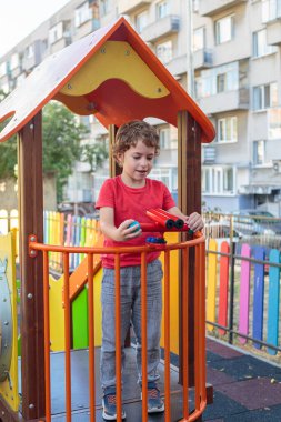 A happy child in a bright red t-shirt stands on a colorful modern playground, enjoying outdoor activities and games on a sunny day