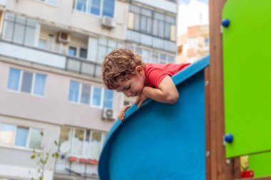 A cheerful child with curly hair laughing while playing and climbing on a bright blue playground structure