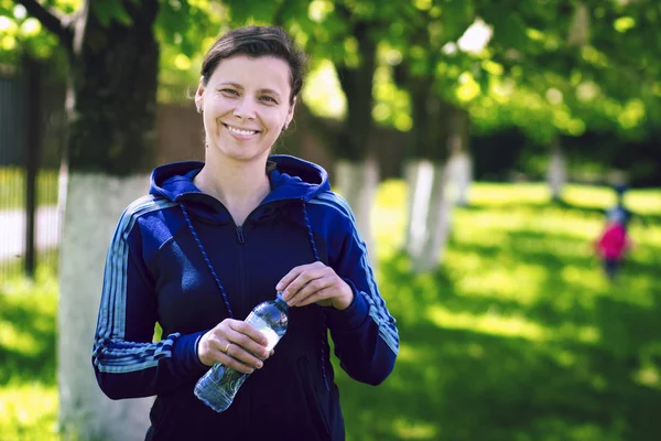 Молодая привлекательная улыбающаяся девушка в спортивной толстовке с бутылкой воды в руке в зеленом летнем парке. Понятие здорового образа жизни — стоковое фото