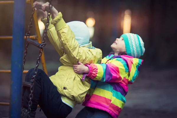 I bambini piccoli giocano in parco giochi in sera in parco su sfondo di luci lucenti . — Foto Stock