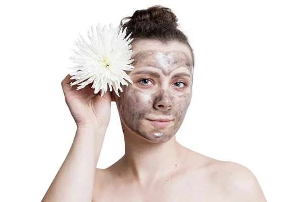 Potret wanita muda Kaukasia dengan topeng wajah kosmetik hitam terisolasi dengan latar belakang putih. Meremajakan kulit wajah. Perawatan Kulit. perawatan spa. Gadis dengan bunga putih di dekat wajahnya. masker wajah . — Stok Foto