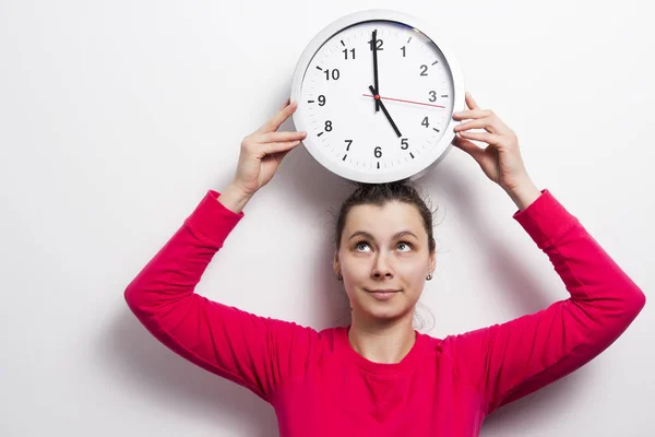 Young woman with clock over her head. Watch the time concept. girl is holding round white clock against white wall background.