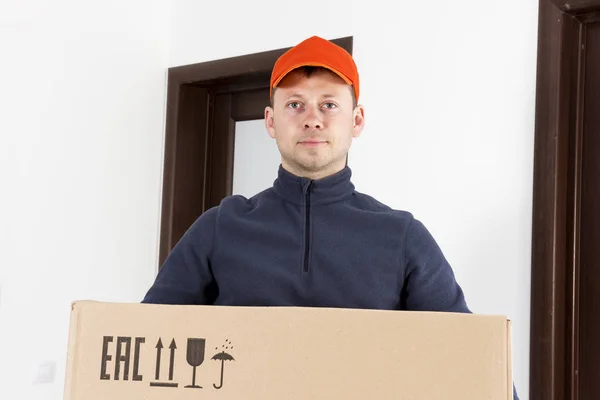 courier in apartment holds cardboard box with fragile load. Delivery of parcel by mail. postman in cap with large cardboard box. delivery service worker with box.