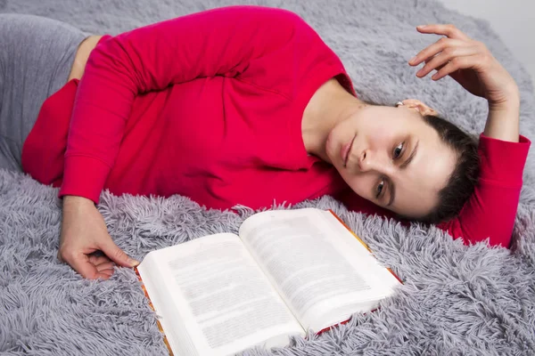 Girl is tired of reading book lying on bed. Attractive young brunette woman lies on bed with book in bedroom. Thinks about meaning of book you read. — Stock Photo, Image