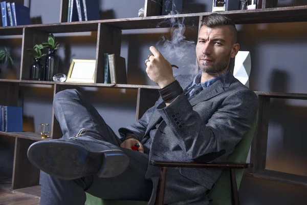 elegant man in suit sits in chair and smokes pipe. handsome stylish rich brutal man in luxurious apartment. Serious view of successful businessman. Clubs of smoke from a cigar in a dark interior.