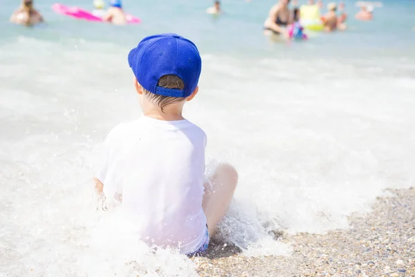 El niño está sentado de espaldas en la playa en un día claro y soleado de verano. niño en la playa tropical en el fondo de las personas bañándose en el mar. Bañarse en agua de mar caliente . — Foto de Stock