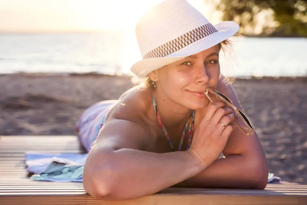 Милий красивий привабливий засмаглий леді в капелюсі лежить на шезлонгу на морському пляжі на заході сонця в теплий літній вечір. Красива жінка посміхається і виглядає сексуально і тримає сонцезахисні окуляри. дівчина ввечері на пляжі . — стокове фото