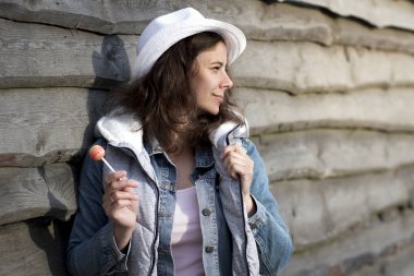 Güzel kız kovboy şapkası ve denim ceket karamelli şeker chupa chups çiftlikte ahşap çit yakınındaki sopa ile. Tatlı çekici Amerikalı kadın portresi yuvarlak Icicle çiftlikte