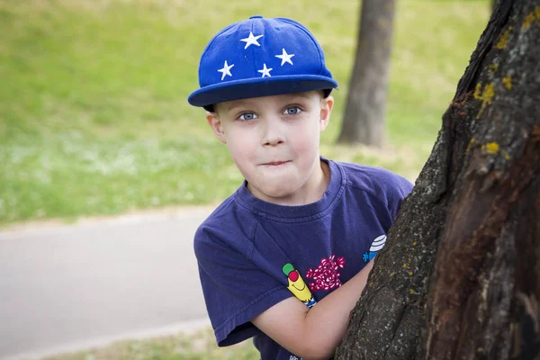 Retrato de un chico caucásico con una gorra asomándose detrás de un árbol . — Foto de Stock