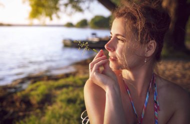 Çekici kız bir yaz akşamı göl kıyısında, günbatımında görünüyor ve ağzına bir saman tutar.