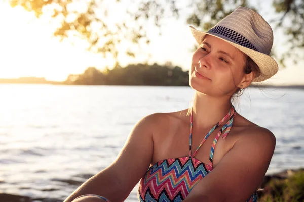 Гарна дівчина в капелюсі сидить на березі озера в теплий літній вечір. Молода жінка за містом на відкритому повітрі . — стокове фото
