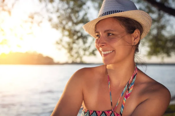 Усміхнений щасливий красивий засмаглий леді в літньому капелюсі на березі озера на тлі заходу сонця над горизонтом. Прекрасна дівчина на пляжі . — стокове фото