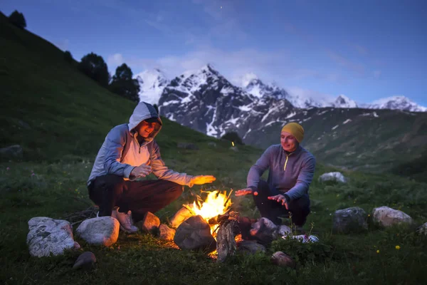 Tourists sit in the evening near fire on background of beautiful mountain landscape. Travelers relax after hike at mountains. Leisure activity.