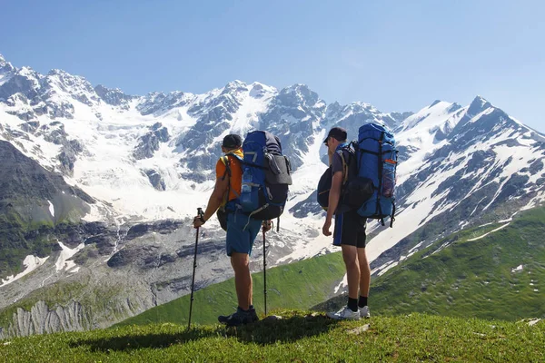 Tourists with hiking backpacks on the background of a rocky snow mountain on a clear summer day. Hiking in mountains with friends. Two travelers in the highlands look at the beautiful mountain scenery
