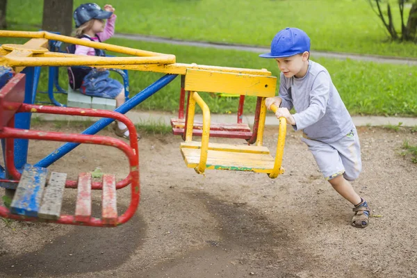 I bambini piccoli rotolano l'un l'altro su caroselli oscillazioni sul parco giochi per bambini nel cortile. Un ragazzo e una ragazza cavalcano sulla giostra. Divertimento per bambini. Bambini sorridenti felici . — Foto Stock