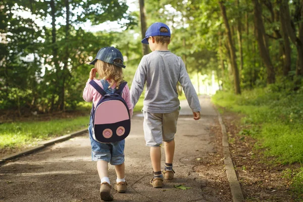 Маленькие друзья дети держатся за руки и ходят по дорожке в парке в летний день. Мальчик и девочка прогуливаются по дорожкам в парке. Детская дружба . — стоковое фото