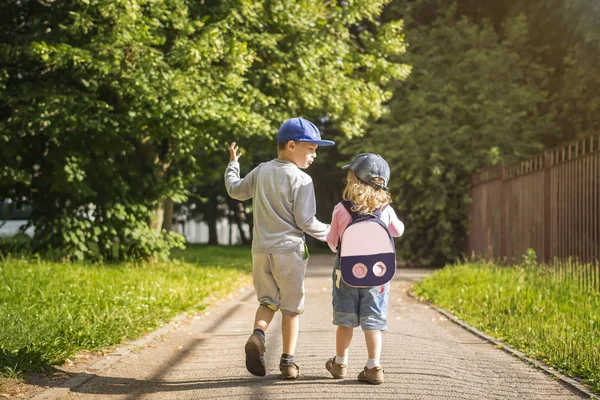 Dua anak kecil teman laki-laki dan perempuan berpegangan tangan dan berjalan di sepanjang jalan di taman musim panas hijau pada sore hari cerah. Persahabatan anak. Anak-anak berjalan di taman — Stok Foto