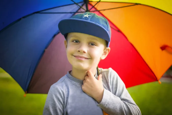 Retrato de un niño blanco en una gorra con un colorido paraguas en un verde parque de verano en un día soleado . — Foto de Stock