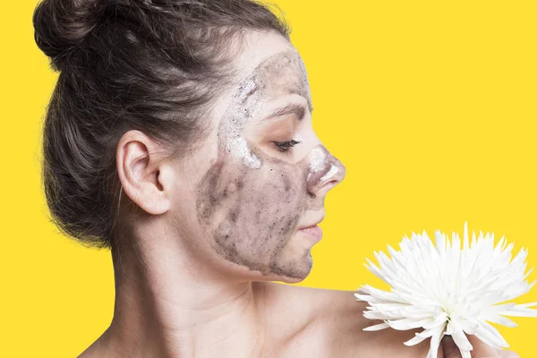 Gezicht van de vrouw in profiel met een cosmetische gezichtsmasker op gele achtergrond met witte bloem. Zachte jonge brunette meisje met een gezichtsmasker — Stockfoto