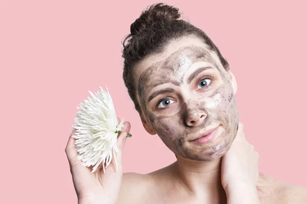 Ung vacker kvinna med ansiktsmask och vit blomma på en rosa bakgrund. Begreppet Ansiktsbehandling hudvård — Stockfoto