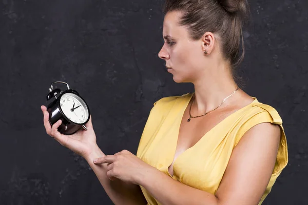 Una giovane donna sta tenendo un orologio. Concetto temporale. La ragazza guarda l'orologio. Studio ritratto di una signora in abito su sfondo nero — Foto Stock