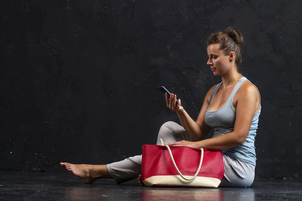 Portrait of a fashionable beautiful young girl with handbag sits and uses smartphone. Studio shot of stylish modern woman after workout yoga. Healthy life style — Stock Photo, Image