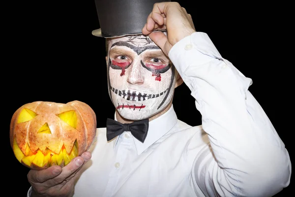 Halloween man with makeup holds yellow pumpkin on black background. Halloween party background. Scary man with a mask of a skeleton in a shirt and hat ready to Halloween celebration.