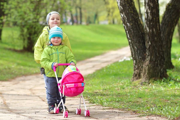 Persahabatan anak-anak. Teman kecil. Anak-anak berjalan di taman hijau. Anak laki-laki dan perempuan dengan mainan kereta untuk boneka. Kakak dan adik bersama-sama — Stok Foto