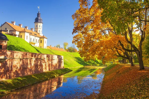 Castello di Nesvizh in autunno soleggiata giornata limpida. Colorati alberi gialli e rossi, cielo blu, erba verde, foglie autunnali che cadono nel fiume nel parco di Nesvizh. Incredibile vista sul famoso castello di Niasvizh, Bielorussia . — Foto Stock