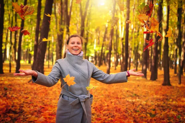 Autumn leaves falling on happy young woman in colourful forest park. Autumnal mood. Fall. Smiling girl in vivid park. Happy woman throwing leaves around on an autumns day.