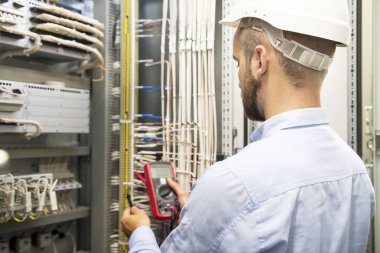 Genç elektrikçi elektrik paneli üzerinde çalışıyor. Elektrikçi mühendisi elektrik tesisatı ve röle koruma sistemi kabloları sınar. Multimetre ile teknisyen sigorta kutusunda. Adam testleri elektrik paneli.