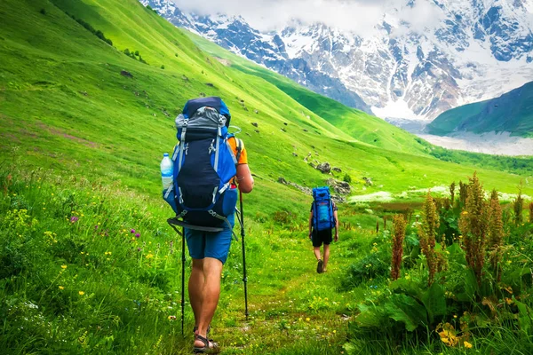 Couple of young travelers hiking with backpacks on beautiful grassy trail in Alps mountains. — Stock Photo, Image