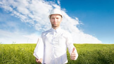 Beyaz miğfer takan inşaatçı yeşil alanda iyi bir ruh hali içinde proje hazırlığında. Projeleri ve planları olan bir işçi. Muhtemel proje.