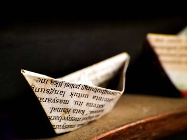 Origami teknesi basılı bir kağıttan yapılmış, görünür bir metni siyah mürekkeple gösteriyor. Tekne ahşap bir yüzeye, daha koyu ahşap tonlarla yerleştiriliyor.. 