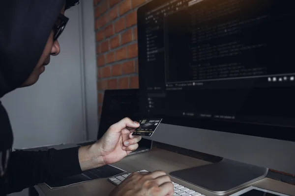 Hacker i hætten arbejder med computer og holder kreditkort - Stock-foto