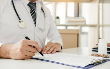 Doktor, elinde kalem tutarak hastanın geçmişini not defterine yazıyor ve hastayla ilaç ve tedavi hakkında konuşuyor..