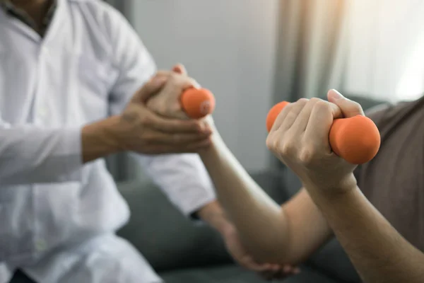 Asian physiotherapist helping a patient lifting dumbbells work through his recovery with weights in clinic room.