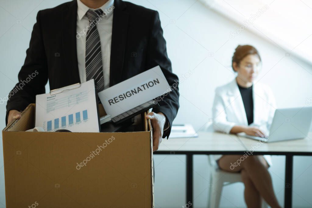 Front view with the male employee standing holding office supplies in the paper box going to submit a resignation letter while a female employee is working behind.