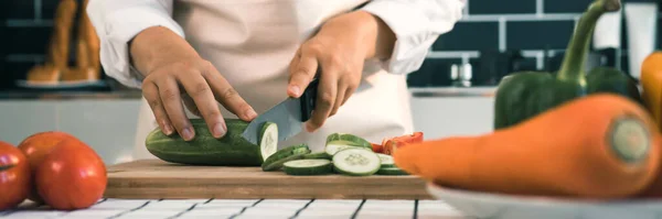 Housewife using knife and hands cutting cucumber on wooden board in kitchen room.