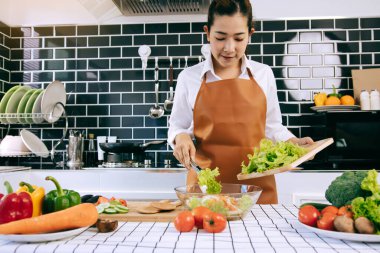 Asyalı ev hanımı salatayı ahşap kesme tahtasından mutfak odasındaki salata kabına götürmek için maşa kullanıyor..