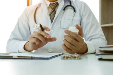 Kendine güveni tam bir doktor elinde bir ilaç şişesi, kıdemli bir hastayla konuşurken yazı yazıyor ve muayenehanede ilaçlarını gözden geçiriyor..