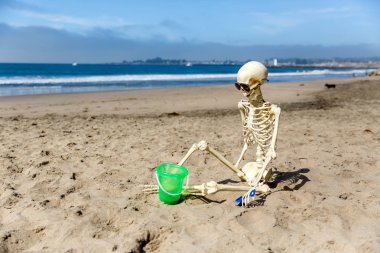 İskelet bir kova ve kürek ile kum da oynarken sahilde oturur