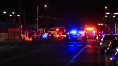 Odaklanamayan olay mahalli. İnsanlar karşıdan karşıya geçiyor. Yanıp sönen polis ve yangın ışıkları önünde yürüyorlar.