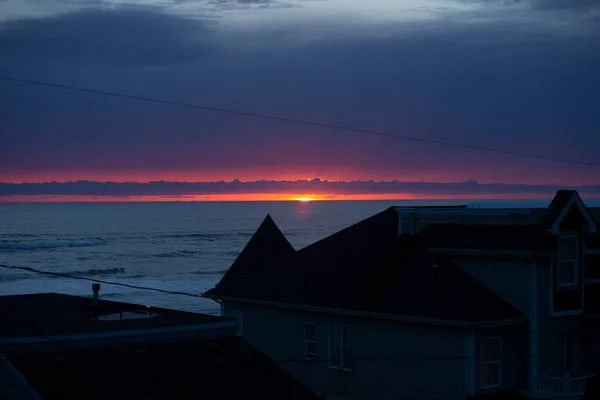 Last light from sunset over the ocean beyond a house and garage roof bisected by power lines