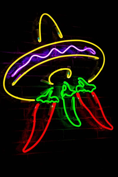 Neon brilhante estilizado sombrero mexicano e pimentas vermelhas e luzes embaçadas no fundo da parede de tijolo preto . — Fotografia de Stock